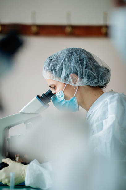 istock Female scientist in protective headwear and mask looking through microscope oculars 1305941134