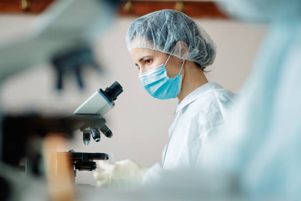 istock Female scientist in protective headwear and mask working in microscopy lab 1305942315