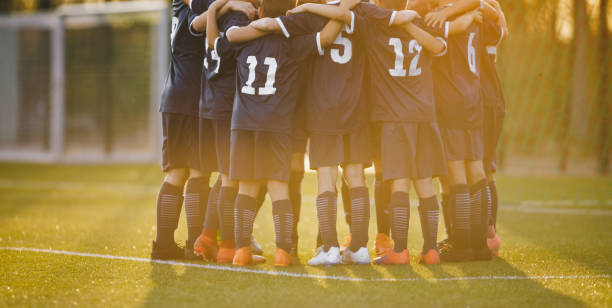 group of children huddling with coach. summer sunset at the stadium in the background. youth soccer football team group photo. happy boys soccer players kicking tournament. school boys in blue jerseys - sports uniform fotos imagens e fotografias de stock