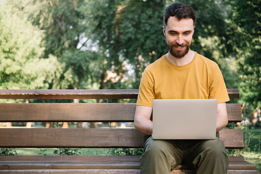Smiling bearded man using laptop computer, shopping online, sitting on bench. Successful freelancer working in park, typing on keyboard
