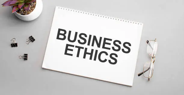 Photo of BUSINESS ETHICS is written in a white notebook next to a pencil, black-framed glasses and a green plant.