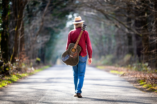 Color image depicting the rear view of a man wearing a red check shirt, denim jeans and a straw hat while walking on a beautiful tree lined country road. He is enjoying the freedom of exploring and wandering in nature, and he has an acoustic guitar strapped to his back. Room for copy space.