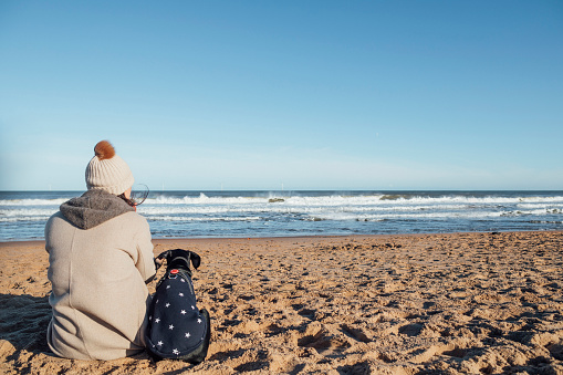 Rear view of a woman walking her pet doberman puppy at the beach in the North East of England. They are taking a break to sit down together side by side.