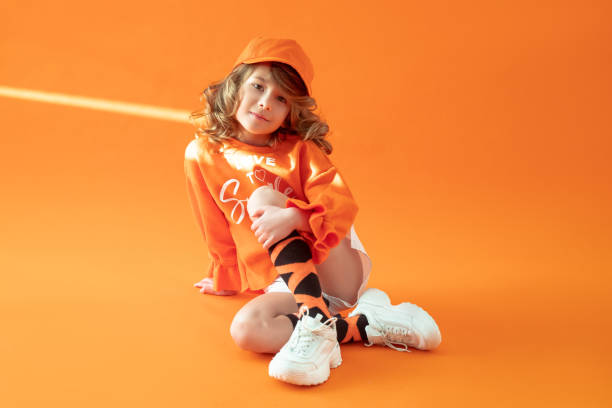 kleines mädchen 6 jahre alt, sitzt auf dem boden im studio auf orangefarbenem hintergrund, posiert in lässiger kleidung, mütze und turnschuhen. kindheit, mode, werbung, sport. nahaufnahme, kopierbereich. sonnenstrahl auf dem kopf - 6 7 years stock-fotos und bilder