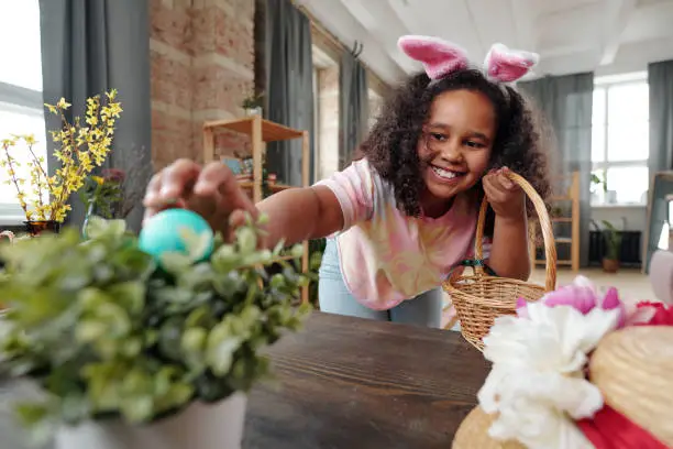 Happy mixed-race little girl with toothy smile bending over wooden table while putting painted Easter egg into flowerpot with domestic plant