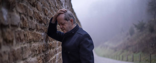 istock While leaning against a worn out wall, a senior man alone in misty forest, lonely and abandoned in gloomy atmospheric mood 1305963418