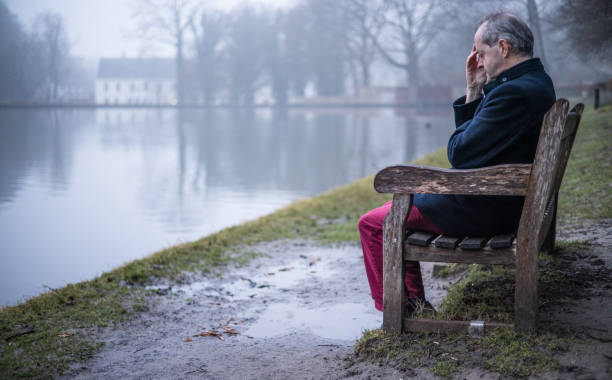 istock Sitting alone on a bench besides a lake, a middle aged man alone in misty forest, lonely and abandoned in gloomy atmospheric mood 1305963647