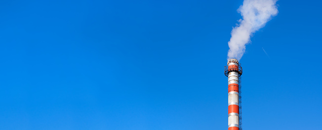 Factory chimney against the clear blue sky. Plane flying in the background. Wide banner with copy space