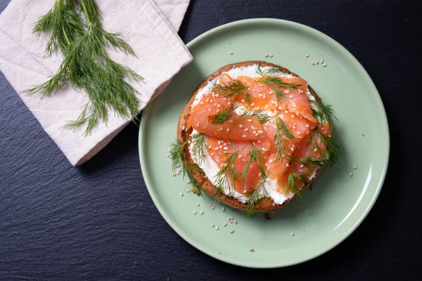 istock half bun oiled with fresh cream, decorated with smoked fish and sprinkled with fresh dill and sesame seeds on a plate, fresh dill and napkin, black background 1305965338