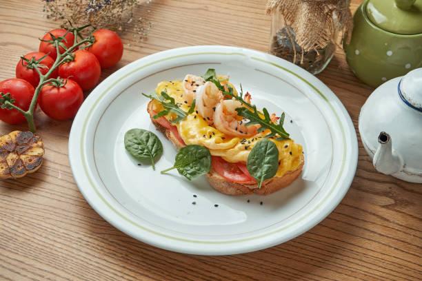 An appetizing and hearty breakfast - a sandwich with scrambled eggs, shrimps, tomatoes and hollandaise sauce, served in a white plate on a wooden background. Restaurant food An appetizing and hearty breakfast - a sandwich with scrambled eggs, shrimps, tomatoes and hollandaise sauce, served in a white plate on a wooden background. Restaurant food egg cherry tomato rye stock pictures, royalty-free photos & images