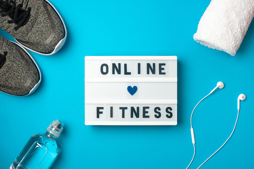Online fitness - text on display lightbox on blue background. Flat lay - Sneakers, a bottle of water, white headphones. Sport concept.