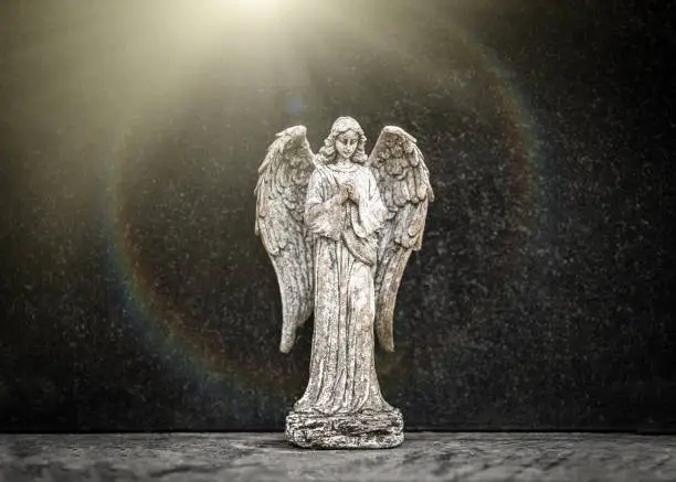 Photo of Small old weathered stone weeping angel with wings ornament standing in sorrow on black granite grave headstone in peaceful churchyard with heavenly sunlight golden rays shining in