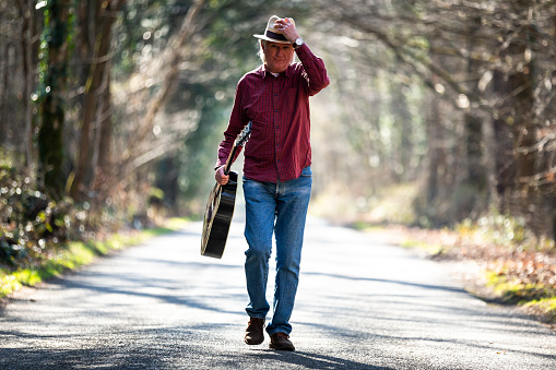 Color image depicting the front view of a man wearing a red check shirt, denim jeans and a straw hat while walking on a beautiful tree lined country road. He is enjoying the freedom of exploring and wandering in nature, and he is carrying a black acoustic guitar. Room for copy space.