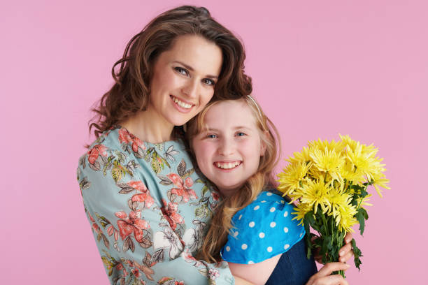 smiling elegant mother and daughter hugging on pink Portrait of smiling elegant mother and daughter with long wavy hair with yellow chrysanthemums flowers hugging isolated on pink background. holding child flower april stock pictures, royalty-free photos & images