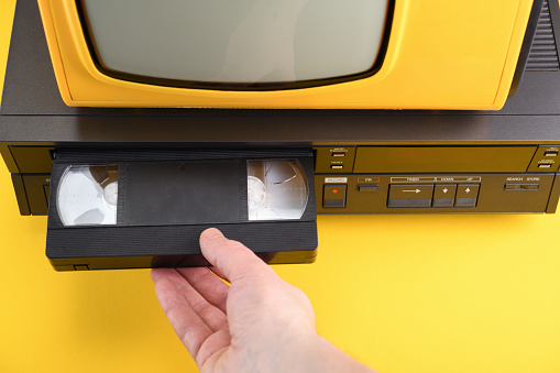 VHS videocassette is put into the video recorder to watch the video. Old yellow vintage TV with VCR and videotape on black background from 1980s, 1990s, 2000s.