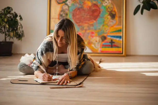 Beautiful active woman sitting in lotus position and banding over to write down her thoughts in notebook after meditation.  She lighted scented stick