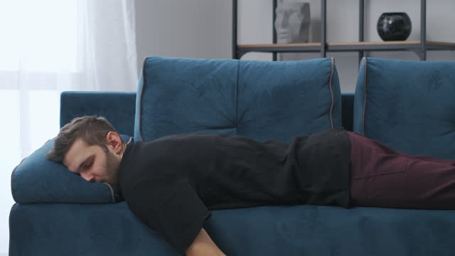 exhausted man is falling on blue couch in apartment, slow motion, adult man is resting at home