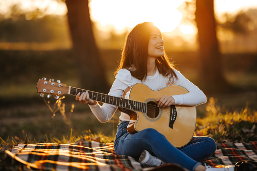 One carefree beautiful young woman with long hair playing acoustic guitar and enjoying picnic day in nature at beautiful sunset in background