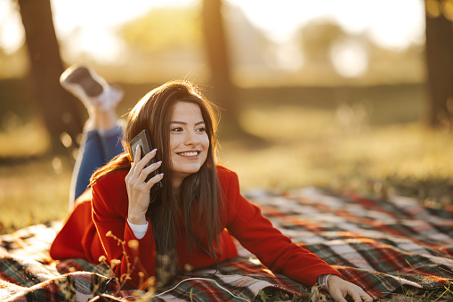 Beautiful young woman lying on picnic blanket and calling friends on mobile phone  in park with beautiful sunset in background