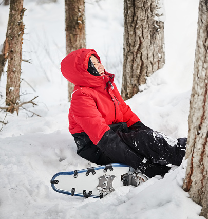 Happy young girl in warm jacket sitting in snowbank while snowshoeing outdoors