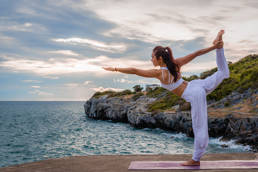 Beautiful Asian woman is practicing yoga on the asana in outdoors with sea and sky background. Healthcare and exercise concept.
