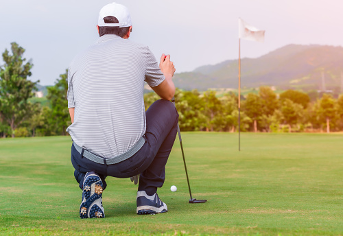 Male golfer kneeling holding golf club and checking path line for golf ball on green.