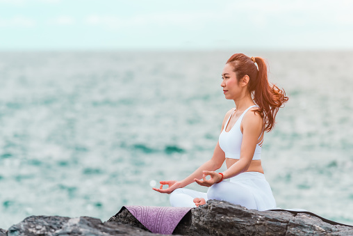 Beautiful Asian woman is practice yoga lotus pose on the rock in outdoors with sea background. Healthcare and exercise concept.