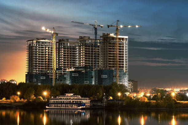 Construction site with new buildings and cranes at evening blue yellow cloudy sky background. Coast of river with  reflections water in front