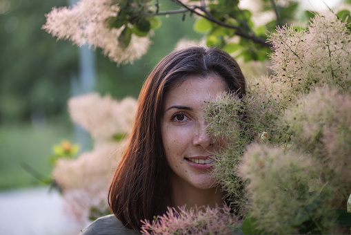 A young woman in a green khaki dress stands by a rosebush Smoky skumpia at sunset in the park. Brunette girl smiling walking in the garden. Close up portrait