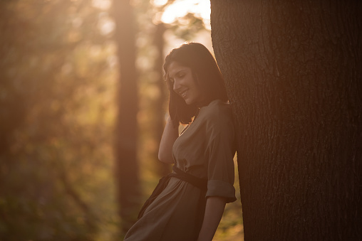 A young woman in a green khaki dress stands with her back against a tree at sunset in the park. Brunette girl smiling, walking in the garden. Close up portrait