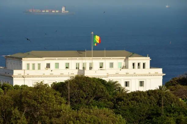 Dakar, Senegal: Senegal's presidential palace with Senegalese flag, the second flag signals that the president is in the palace - Palace of the Republic, residence of the President of the Republic, a historic manor in the Plateau district. Designed by Henri Deglane, completed in 1907, this colonial palace used to be the official residence of the Governor General of French West Africa - Atlantic Ocean and container-ship in the background - Avenue du President Léopold Sédar Senghor.