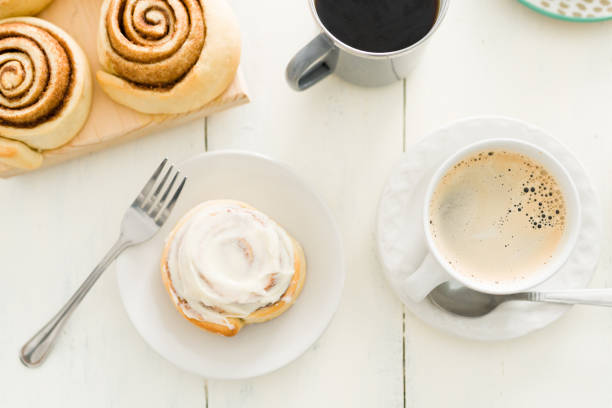 Dessert is served. Flat lay of a glazed cinnamon roll on a white plate next to a hot cup of coffee placed on a wooden kitchen table