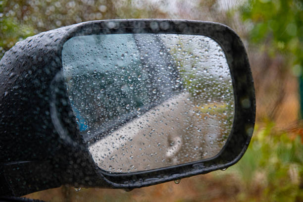 Wet glass texture with raindrops rolling on window. Car side view mirror with defocused reflections and blurred background outside car of autumn colors nature