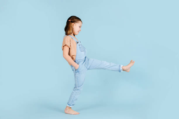 Playful little girl doing wide steps with straight legs over blue background. Playful barefoot little girl doing wide steps with straight legs over blue background.. She has two ponytails, wears overalls. Hands in pockets. Barefoot stock pictures, royalty-free photos & images