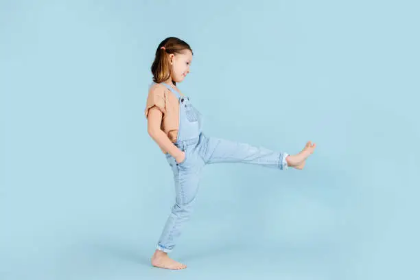 Playful barefoot little girl doing wide steps with straight legs over blue background.. She has two ponytails, wears overalls. Hands in pockets.
