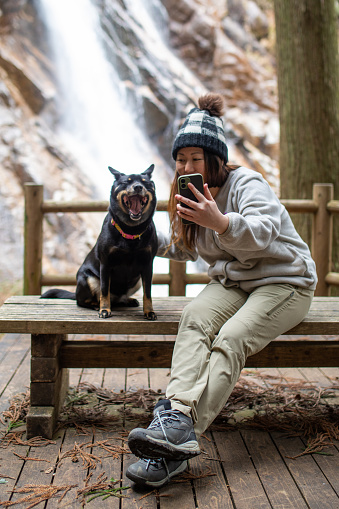 A Japanese woman in warm clothing is having fun taking a selfie with her dog who has its mouth wide open in the outdoors by a waterfall.