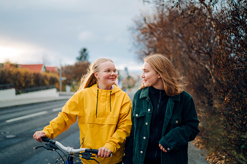 Teenage friends walking down the street with a bike. Two girls smiling at each other while walking outdoors on a street with a bicycle outdoors.