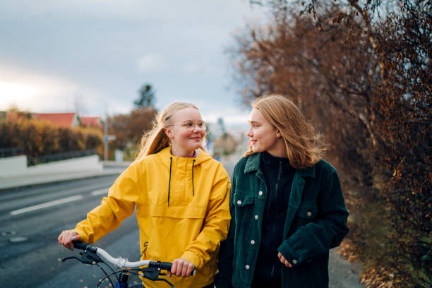 Teenage girls walking outdoors with a bike Teenage friends walking down the street with a bike. Two girls smiling at each other while walking outdoors on a street with a bicycle outdoors. 15 year old blonde girl stock pictures, royalty-free photos & images