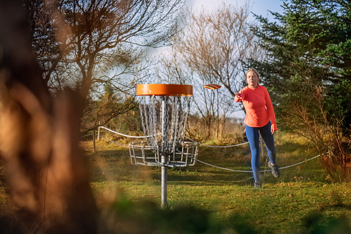 Woman tossing a disc into the basket goal. Caucasian woman playing disc golf game outdoors in a park.