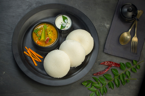 Idly or Idli, south indian main breakfast item which is beautifully arranged in a black plate with grey coloured texture with kitchen background.