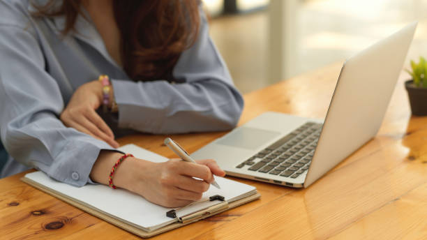 istock Female office worker hand with pen writing on paperwork while working with laptop on wooden table 1306093639