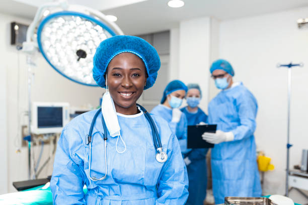 手術室でバックグラウンドで演奏する同僚とカメラを見ているアフリカ系アメリカ人の外科医女性のクローズアップ。医学の概念 - 麻酔医 ストックフォトと画像