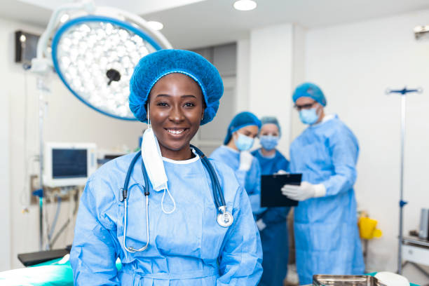 Close-up of a African American surgeon woman looking at camera with colleagues performing in background in operation room. The concept of medicine Close-up of a African American surgeon woman looking at camera with colleagues performing in background in operation room. The concept of medicine anaesthetist stock pictures, royalty-free photos & images