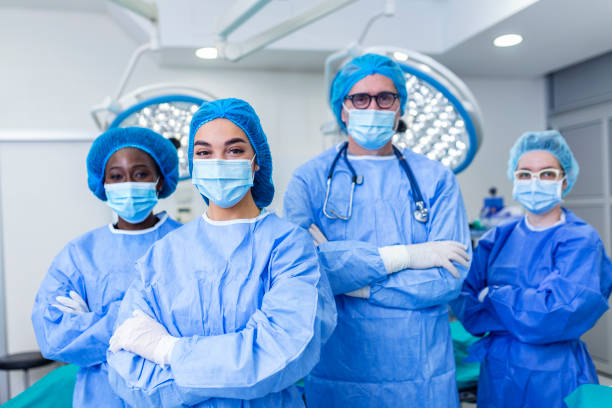 multi-ethnic group of four healthcare workers, a team of doctors, surgeons and nurses, performing surgery on a patient in a hospital operating room. They are looking at the camera. multi-ethnic group of four healthcare workers, a team of doctors, surgeons and nurses, performing surgery on a patient in a hospital operating room. They are looking at the camera. anaesthetist stock pictures, royalty-free photos & images