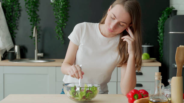 unhappy woman eating vegetable salad at table in kitchen. displeased young woman eating green leaf lettuce - anorexia imagens e fotografias de stock