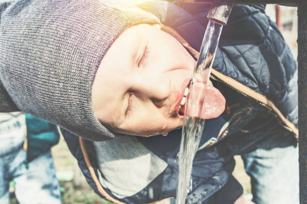Funny little boy in grey knitted hat drinks water from vintage water pump in village house yard on sunny day close view