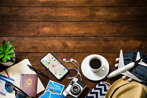 Top view of various traveling stuff such as an opened notebook, a coffee cup, a camera with some snapshots, a smartphone with a map on the screen, a tiny airplane, two passports with boarding passes and some summer clothing like a hat and sunglasses. All the objects are at the lower side of the image leaving a useful copy space at the top on a rustic wooden desk.  Studio shot taken with Canon EOS 6D Mark II and Canon EF 24-105 mm f/4L