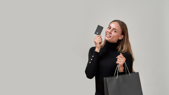 Joyful young woman wearing black clothes smiling at camera, showing plastic credit card while posing with shopping bag isolated over gray background. Fashion, banking and payment concept