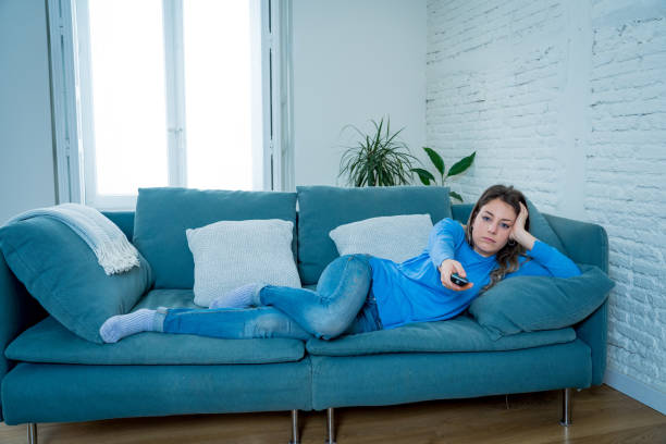 Bored woman watching TV helpless in self Isolation at home during mandatory lockdown due to coronavirus outbreak. Young upset woman on sofa using control remote zapping bored of TV and sedentary life. mujer aburrida viendo la televisión indefensa en auto aislamiento en casa durante el encierro obligatorio debido al brote de coronavirus. joven mujer molesta en el sofá usando control remoto zapping aburrido de la televisión y la vida sedentaria.