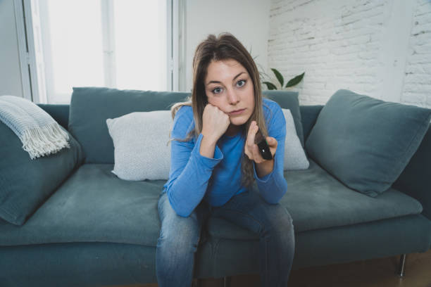 Bored woman watching TV helpless in self Isolation at home during mandatory lockdown due to coronavirus outbreak. Young upset woman on sofa using control remote zapping bored of TV and sedentary life. mujer aburrida viendo la televisión indefensa en auto aislamiento en casa durante el encierro obligatorio debido al brote de coronavirus. joven mujer molesta en el sofá usando control remoto zapping aburrido de la televisión y la vida sedentaria.